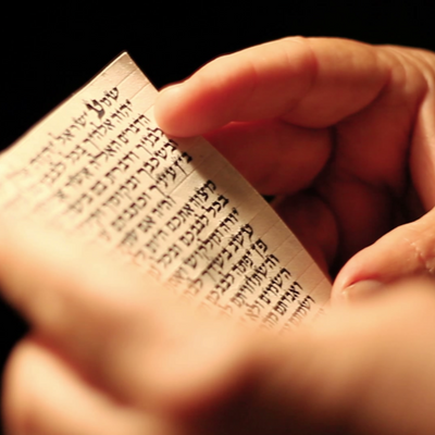 ידיים מחזיקות קלף מזוזה קטן וכתוב, ברקע מטושטש ובתאורה חמימה.
