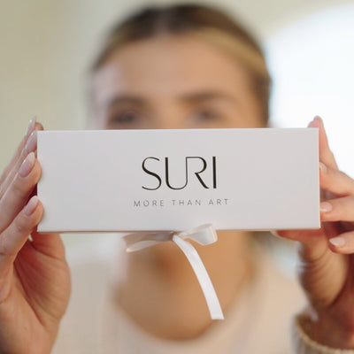 אישה מחזיקה קופסת מתנה לבנה של SURI לבית מזוזה וקלף, עם סרט וברכת תלייה, "MORE THAN ART" על הקופסה.
